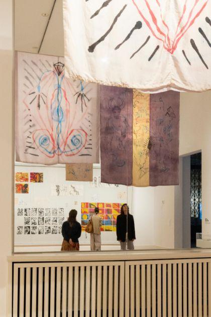 Menschen betrachten Kunstwerke mit Stoffbannern und Wandzeichnungen in einer Ausstellung.