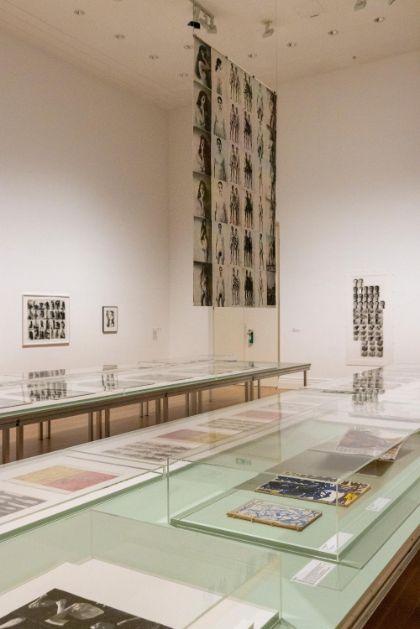 Museumsausstellung mit Glastischen, auf denen Kunstwerke und Fotografien gezeigt werden. Große Wand mit Kunstwerken im Hintergrund.