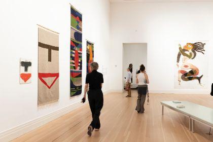 Personen betrachten in einer Galerie abstrakte Kunst, darunter Wandteppiche und ein Gemälde.