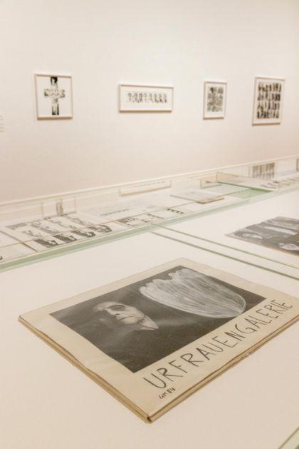 Ausstellungsraum mit gerahmten Bildern an der Wand und Informationsmaterialien in Glasvitrinen.