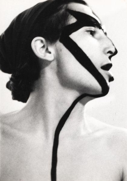 A black‑and‑white profile portrait of a person with the head slightly tilted upward. Wide, dark paint stripes run diagonally across the face and down the neck, crossing the forehead, eye area, and cheek.
