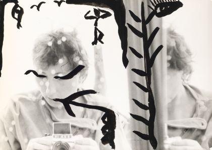 Ein mit schwarzen Mustern bemaltes Selbstporträt von Gabriele Stötzer, die sich mit einer Analogkamera im Spiegel fotografiert.