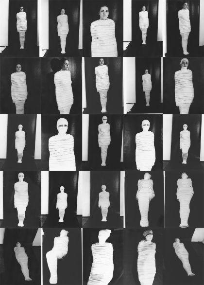 An arrangement of multiple black-and-white photographs displayed in a regular grid. Each photograph shows a standing or slightly moving human figure completely wrapped in light-colored fabric. The figure appears at varying distances from the camera and in slightly different positions against a dark background.
