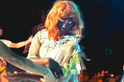 An individual in a light, textured garment with ruffled detailing stands in a dimly lit performance setting. The person holds or touches a pale sculptural arm or prop extending into the frame from the left.