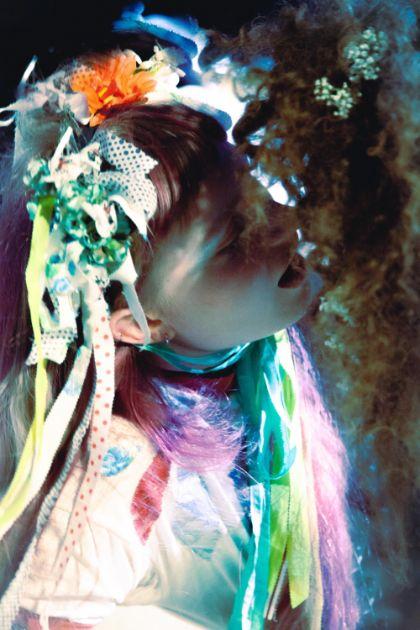 One person is positioned close to the face of another person. The person wears a garment made of light-colored fabric with multicolored decorative elements, including ribbons, patterned fabric pieces, and small floral components arranged around the head. Colorful fabric strips hang down, and the lighting creates strong contrasts of blue, green, and warm tones in the image.