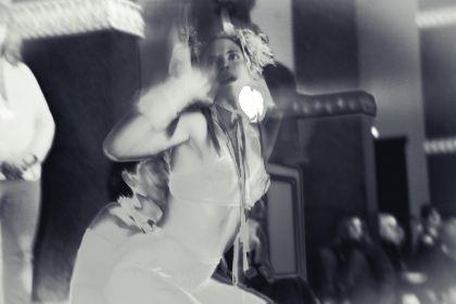 An individual in a light, form‑fitting costume with decorative elements is in a dynamic pose on a stage. Behind the person in the foreground, another person is visible in motion. The scene is presented in high‑contrast black and white, and blurred audience members can be seen in the background.