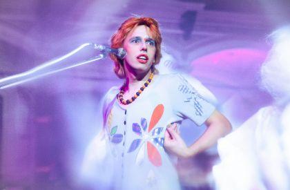 An individual stands on a stage wearing a light-colored garment decorated with large, colorful shapes resembling petals or abstract motifs. A beaded necklace is visible around the neck. A microphone extends toward the person from the left side of the image. The lighting creates soft purple and pink tones with motion blur around the figure and background.