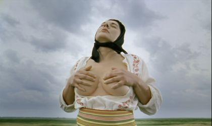 A person wearing a black headscarf, is standing outdoors and is massaging her naked breasts.