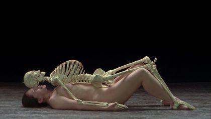 A nude person lies flat on the floor in a side view. The legs are slightly bent, and the arms rest alongside the body. A human skeleton lies on top of the person, mirroring the exact same position.