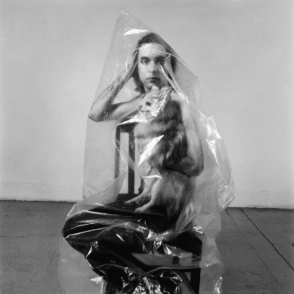 A person is sitting on a chair, resting their arm on the backrest and looking into the camera. A dog is sitting on their lap. A transparent plastic bag is draped over them.