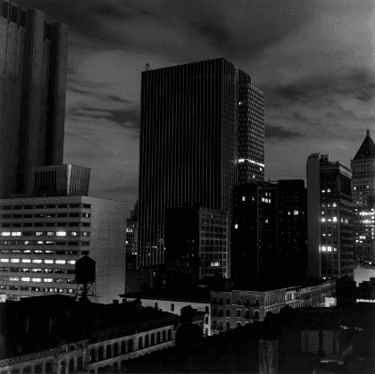 Night view of a city taken from an angled perspective, showing tall buildings. Some windows are illuminated. Clouds move across the sky.