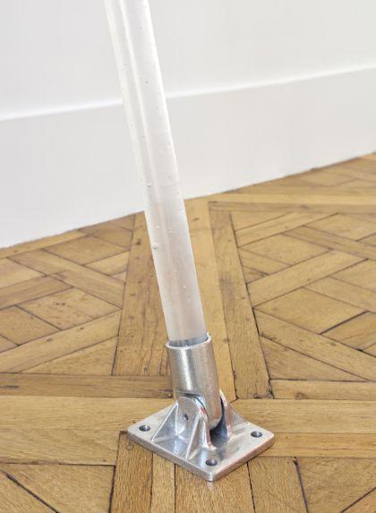 Close-up of a silver hinge on light brown wooden floorboards, with a milky translucent rod extending vertically upward. The construction tilts slightly to one side. A white wall is visible in the background.