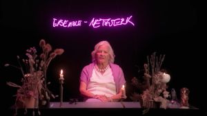 Eine weiblich gelesene, ältere Person mit weißem Haar sitzt zwischen Trockenblumen und brennenden Kerzen. In leuchtenden, pinken Lettern steht an der Wand hinter ihr ENSEMBLE-NETZWERK geschrieben.