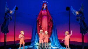 Four people in salmon pink costumes and with a mask over their heads stand in front of a huge statue of the Virgin Mary flanked by two angels.