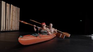 Two people sit in a canoe on a dark stage. Their canoe is in front of a wooden fence.