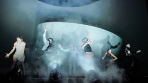 Six people dance expressively in stage fog in front of a white spiral stage set.