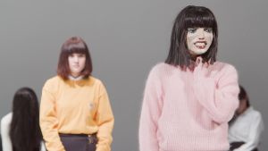 A person in a pink jumper with a mask on their face is looking forwards. A second person in a yellow jumper stands behind her and watches her.