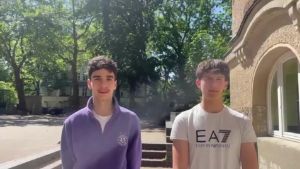 Two people are standing outside on a sunny day. The person on the left is wearing a long-sleeved, purple-coloured top, while the person on the right is wearing a white T-shirt with the words ‘EA7 Emporio Armani’. Green trees, a multi-storey building and a curved façade with stairs can be seen in the background.