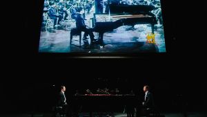 Zwei Pianisten sitzen sich an zwei Klavieren gegenüber. Im Hintergrund befindet sich eine große Leinwand, auf der ebenfalls ein Pianist zu sehen ist, der an einem Flügel sitzt, vor einem Orchester, das ein Dirigent anleitet.