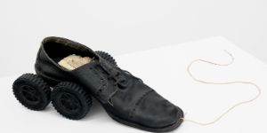 Ein gebrauchter Lederschnürschuh auf einem weißen Tisch, dessen Absatz auf zwei schwarze Räder gestützt ist. Vorne am Schuh ist eine Schnur befestigt.