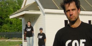 Christoph Schlingensief steht vor einer weißen, aus Holz gefertigten Kapelle. Hinter ihm stehen zwei Personen, sie alle tragen T-Shirts mit dem Schriftzug „COF“.