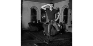 Eine Schwarz-Weiß-Aufnahme von einer Person in einem Raum mit vier Fenstern, Sesseln und Teppichen, die in die Luft springt und dabei eine Hand an die Hüfte und eine Hand an den Kopf hält.