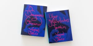 Zwei lilafarbene Bücher liegen auf hellen Hintergrund, auf ihnen steht in magenterfarbener Schrift gedruck "Über gefährliches Terrain. Vaginal Davis und "On Dangerous Ground. Vaginal Davis".