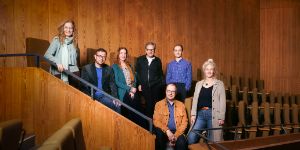 The seven Theatertreffen jury members stand or sit in the balcony of a theatre and look into the camera.