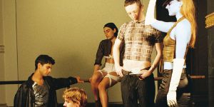 Fünf Performer*innen von Alex Baczyński-Jenkins "Malign Junction (Goodbye, Berlin)" stehen und sitzen auf einem Holzpodest. Sie blicken nachdenklich in unterschiedliche Richtungen.