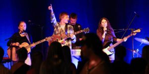 Four young musicians perform on a blue-lit stage with guitars and microphones.