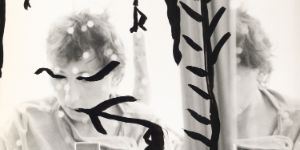 Ein mit schwarzen Mustern bemaltes Selbstporträt von Gabriele Stötzer, die sich mit einer Analogkamera im Spiegel fotografiert.