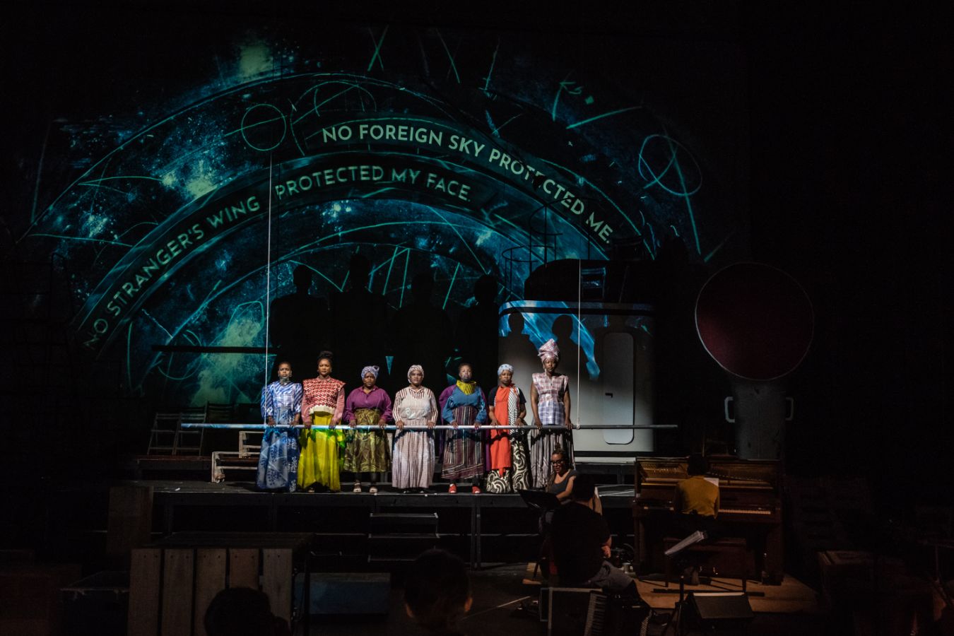 Ein Chor mit Frauen of Colour in bunten Kleidern stehen auf der Bühne. Sie halten sich an einer horizontalen Stange fest.