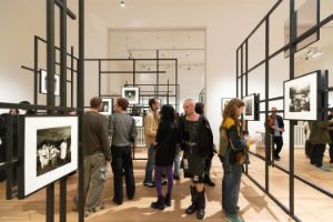 Ein schwarzes Gittersystem mit Schwarz-Weiß-Fotografien der Fotografin Diane Arbus in den Ausstellungsräumen des Gropius Bau. Zwischen den Stellagen sind Besuchende, die sich die Fotografien anschauen.