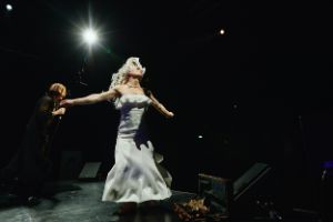 Stage situation: A person wearing a flowing white wig, a white evening gown, and with accentuated arm muscles stretches out their arms and tilts their head back slightly, as if in an emotional moment.