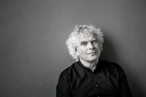 Portrait of a man with grey curly hair  and a black shirt in front of a grey background.