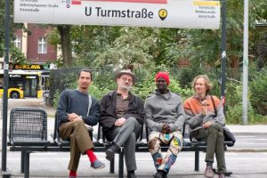 Simon Sieger, Michael Griener, Assane Seck, Joel Grip sitzen auf einer Bank unter dem Schild der U-Bahn-Station "U Turmstraße"