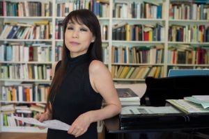 Eine Frau lehnt vor einer Bücherwand am Klavierflügel und schaut in die Kamera.