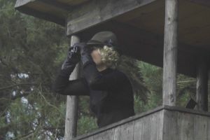 Ein blonde Frau beobachtet auf einem Hochsitz im Wald etwas mit einem Fernglas.