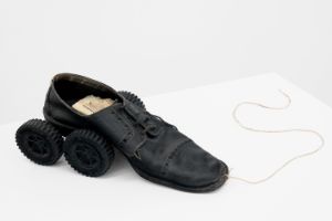A worn lace-up leather shoe on a white table, its heel supported by two black wheels. A piece of string is attatche to the front of the shoe.