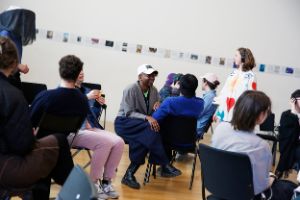 A group is sitting on chairs and looking in different directions. A person standing is wearing headphones and watching a person wearing a hat with a black veil.