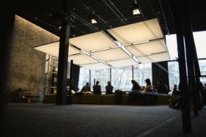 Menschen sitzen auf Sofas im oberen Foyer des Haus der Berliner Festspiele, über ihnen hängt eine aus quadratischen Flächen bestehende Licht-Raum-Installation.