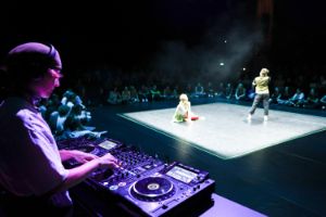 Menschen tanzen auf einer beleuchteten Bühne in einem sonst dunklen im Raum. Im Vordergrund ist angeschnitten ein*e DJ an Turntables zu sehen.