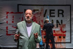 Ein alter Mann erklärt etwas mit erstem Gesicht, im Hintergrund bearbeitet jemand ein blutverschmiertes Plakat