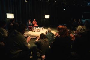 Stage situation: The audience sits in a circle around a round carpet on which there is a flat table with vessels. Two performers sit in front of it, their hands in a gesture of greeting.