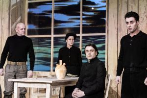 Four people in old-fashioned-looking black clothes are standing in a kitchen table scene where everything is made of wood.