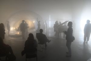 The silhouettes of several standing or seated people are recognisable in a dimly lit, misty room containing a transparent sphere and a grand piano.