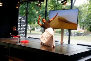 A dancer wearing a black T-shirt, white skirt, and high heels makes sweeping gestures in front of a screen showing a desert landscape.