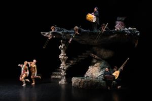 On stage there is a two-storey rock formation. Musicians are scattered across it and two people in colourful costumes are dancing next to it.