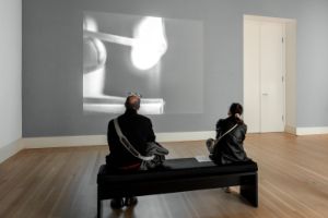 Ein Video von einer Person mit einem Streichholz wird in einem Ausstellungsraum an eine Wand projiziert, davor sitzen zwei Personen auf einer Bank.