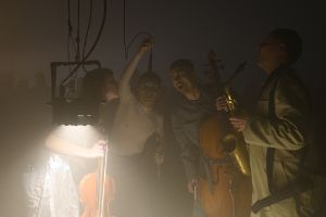 Four people holding instruments in their hands stand in a dark room around a microphone. The silhouettes of other people are recognisable in the background.